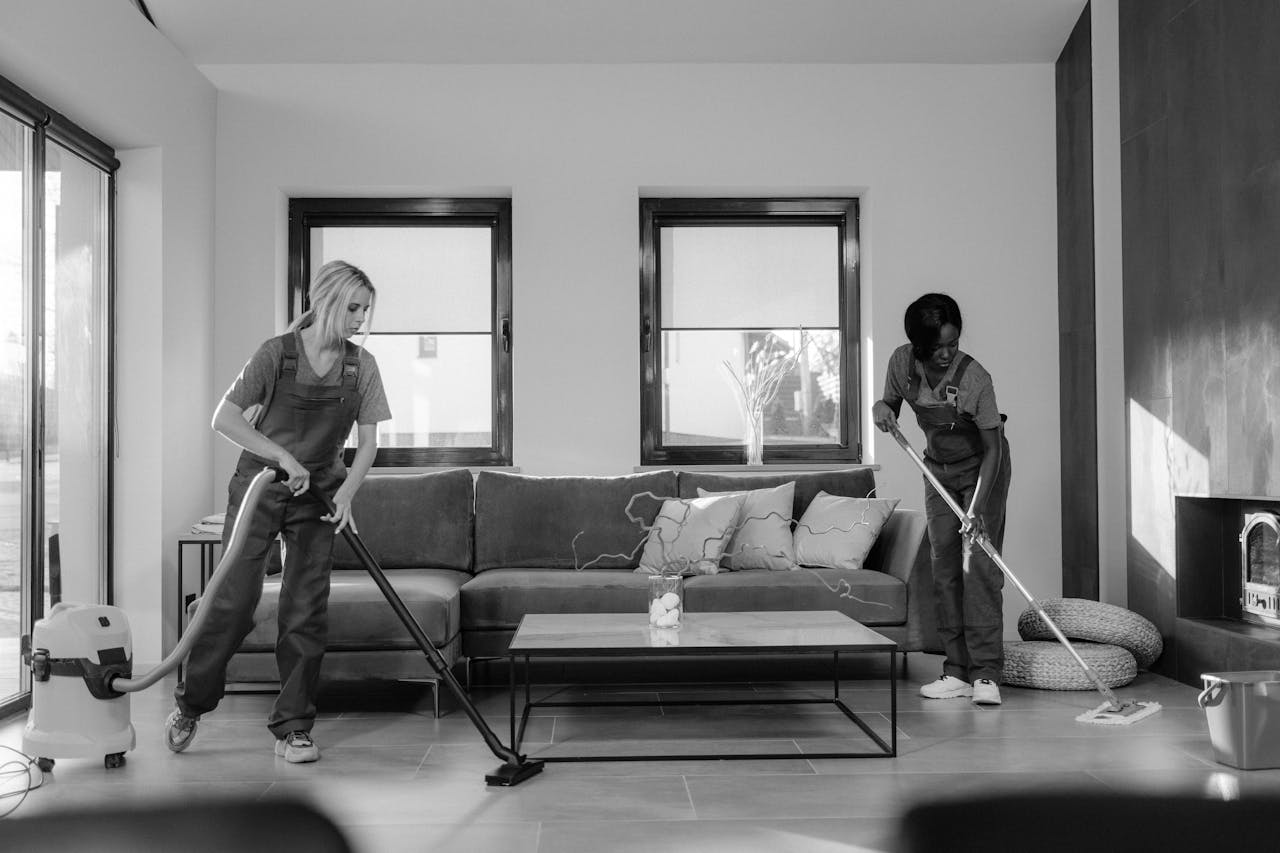 heros-img Two women in work clothes cleaning a modern living room using a vacuum and mop.